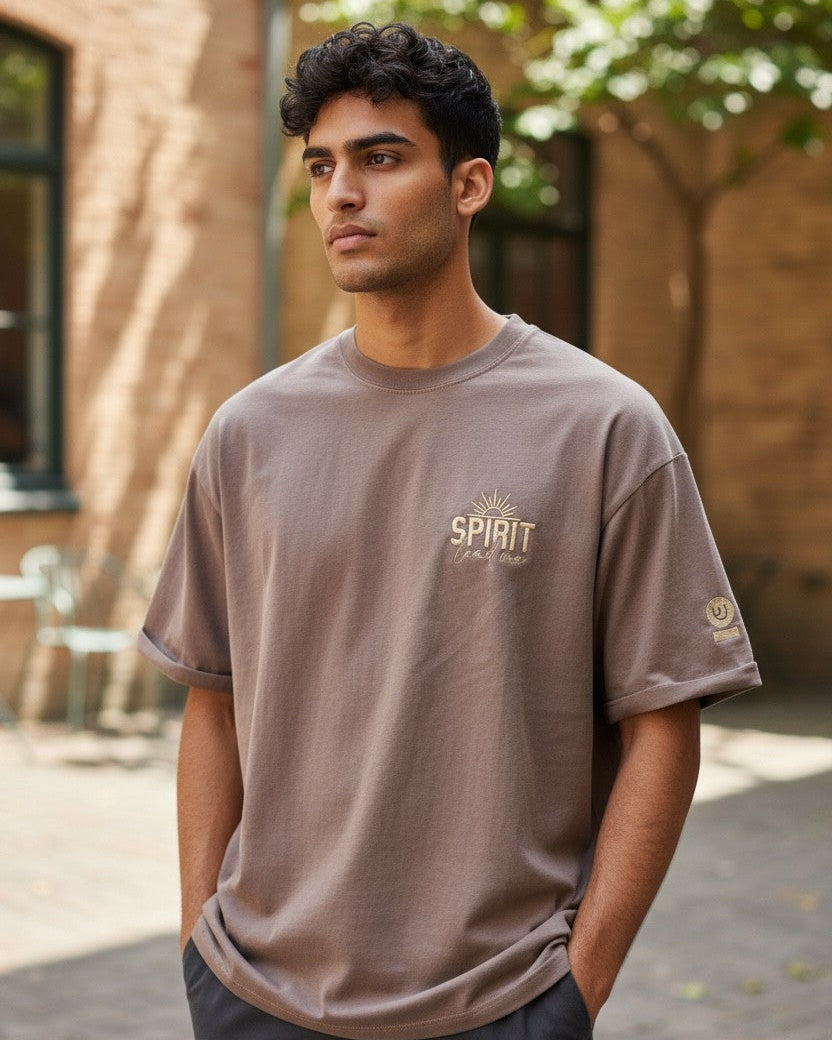 Man wearing a brown t-shirt with text standing outdoors.