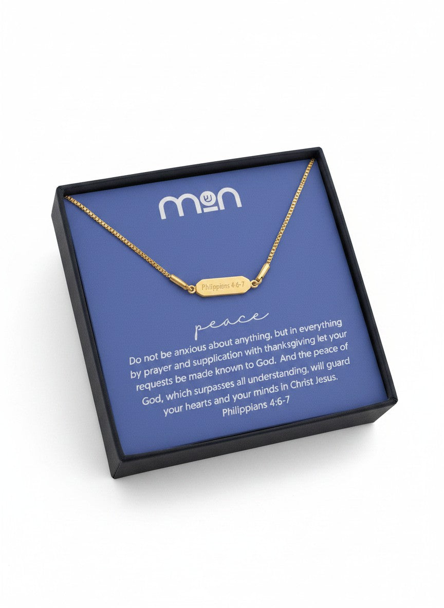 Gold necklace with 'Philippians 4:6-7' on the back of the pendant, placed on a blue card with 'mon' branding in a black box