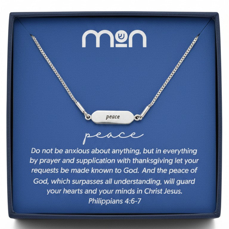 Silver necklace with 'Peace' on the front of the pendant, placed on a blue card with 'mon' branding in a black box