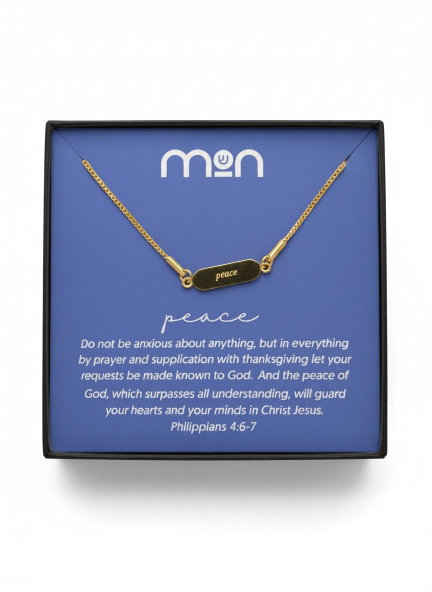 Gold necklace with 'peace' on the front of the pendant, placed on a blue card with 'mon' branding in a black box