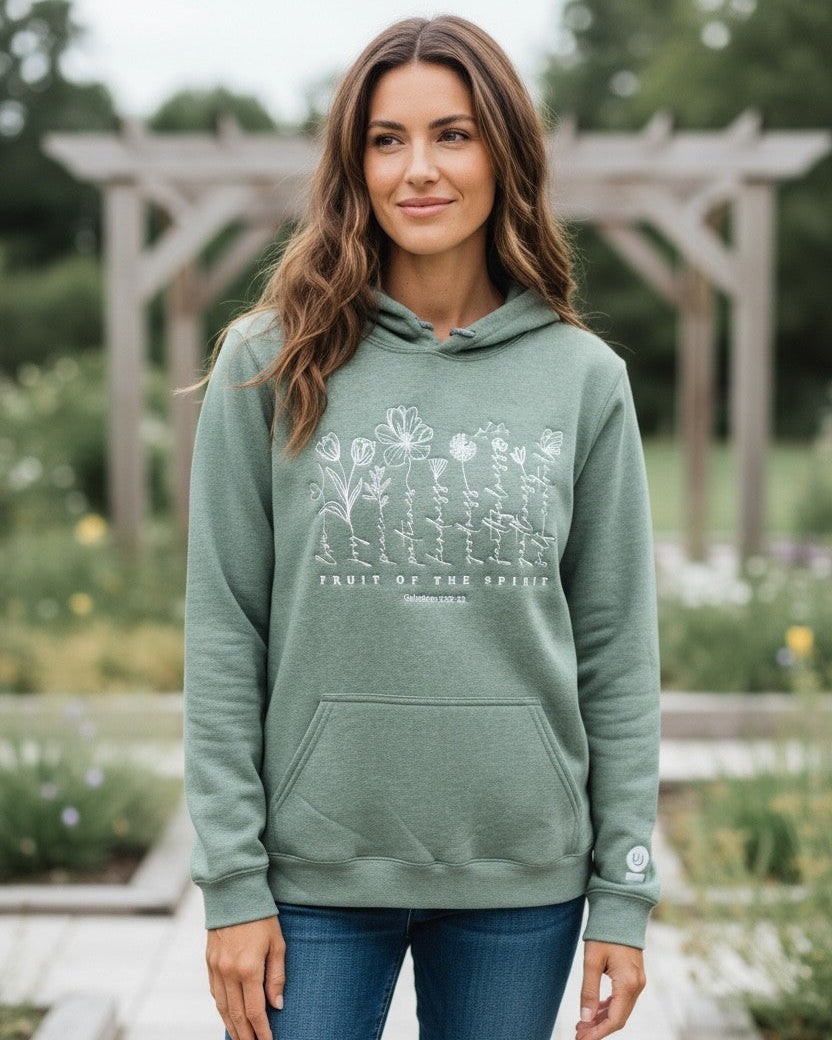 Woman wearing a green hoodie with floral designs and text, standing in a garden.